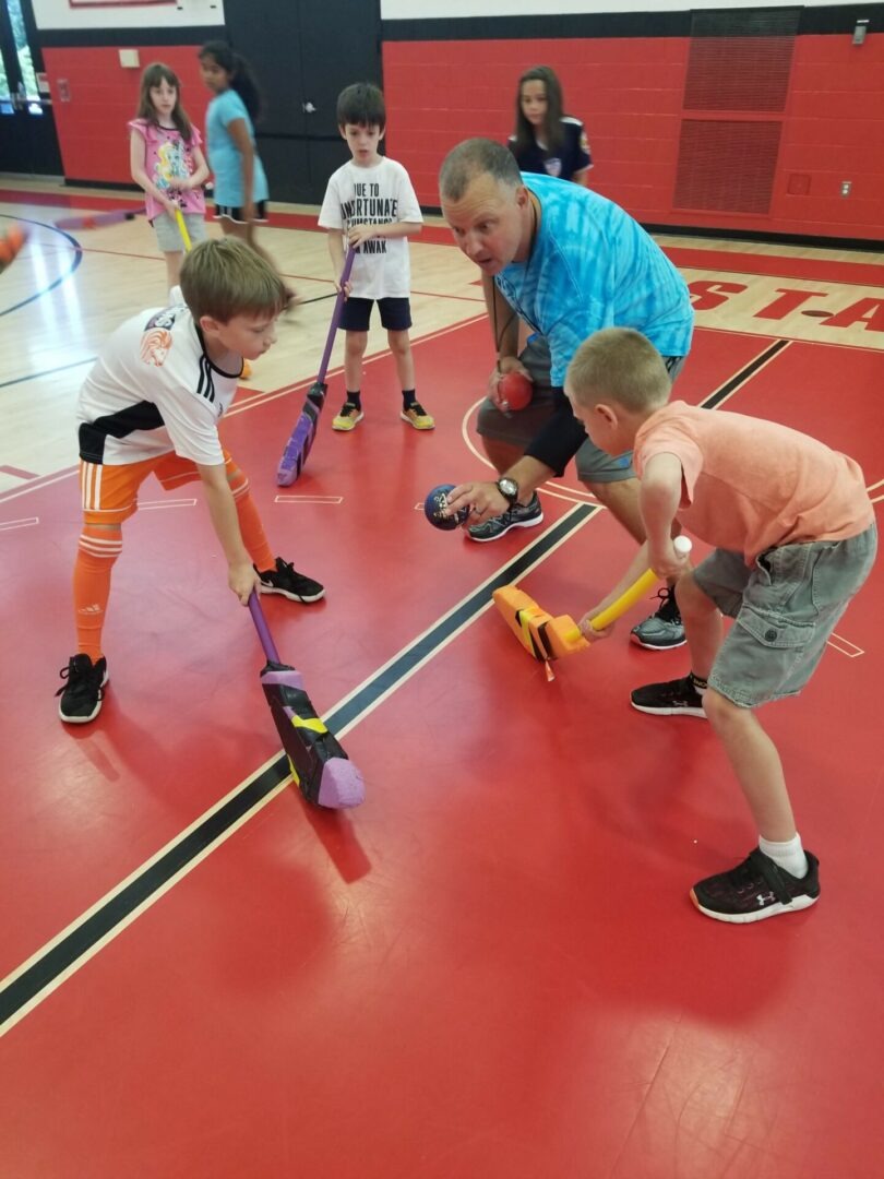 Children engage in a playful sports lesson, using colorful equipment under the guidance of an adult. The setting is a gymnasium.