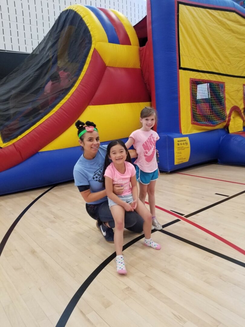A smiling adult holds a girl on her lap while a second girl stands beside them, all in front of an inflatable bounce house in a gym.