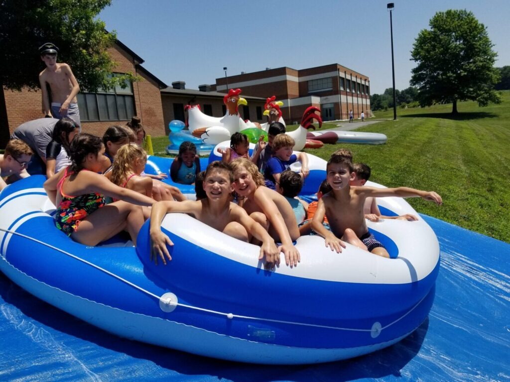 Children joyfully play on a large inflatable raft in a sunny outdoor setting, surrounded by various water attractions and a grassy area.