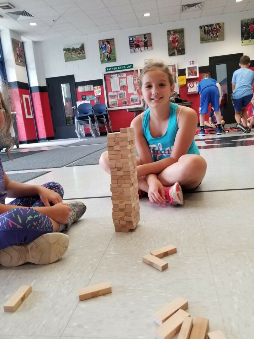 A girl smiles beside a tall stack of wooden blocks on the floor, while a friend sits close by, showcasing teamwork and play in a gym setting.
