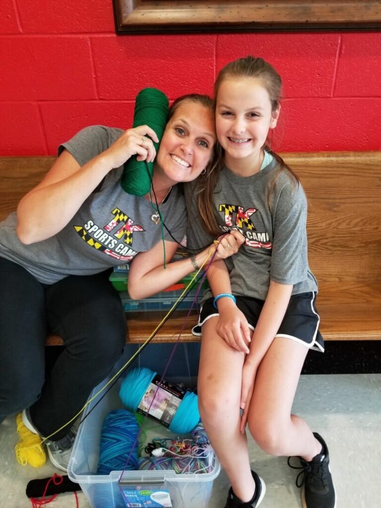 Smiling camp leader and girl pose with yarn, showcasing a fun crafting activity at a sports camp, emphasizing creativity and teamwork.