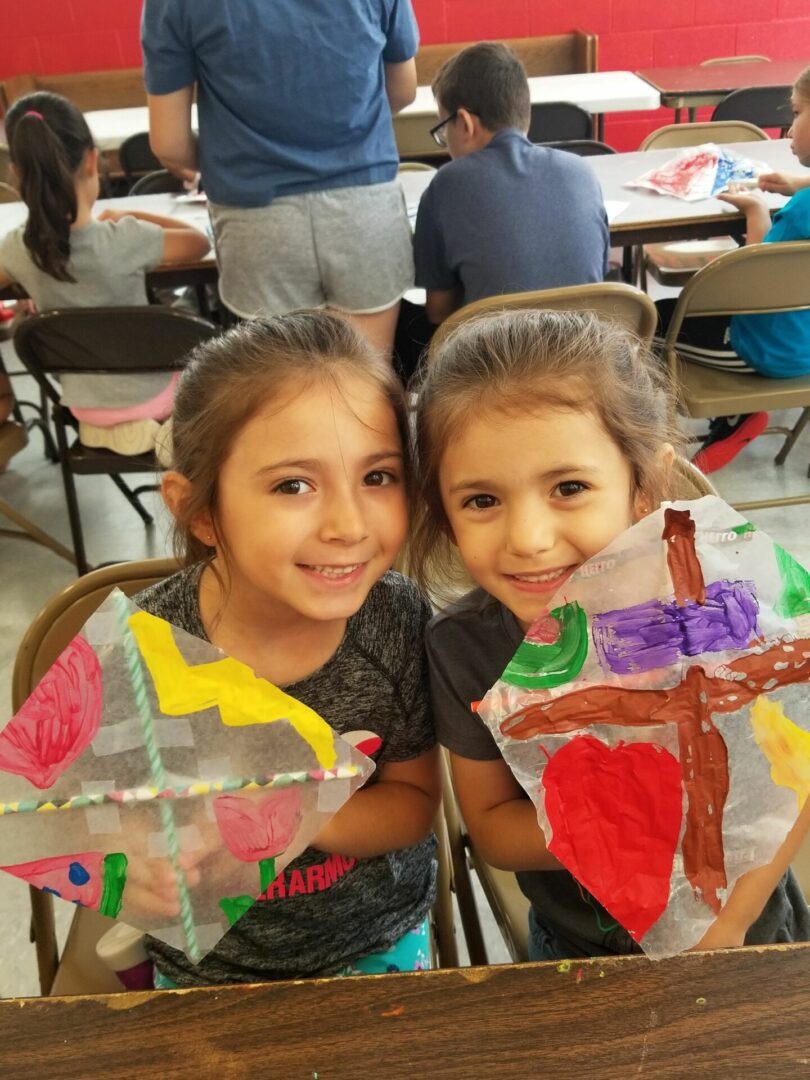 Two young girls smile while holding colorful, hand-painted kites, showcasing their crafts during an art activity in a classroom setting.
