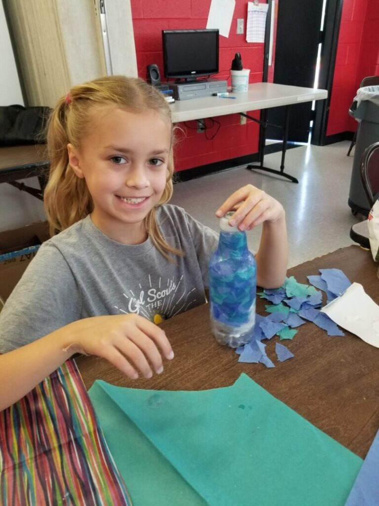 A young girl smiles while holding a decorated plastic bottle made from colorful paper pieces. Craft materials and a workspace are visible in the background.