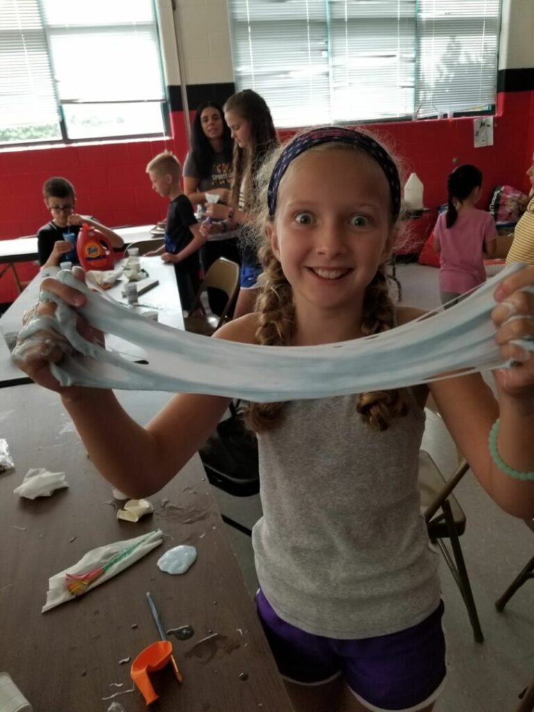 A girl with wide eyes smiles while holding a long piece of blue slime. Other children engage in crafting activities in the background.