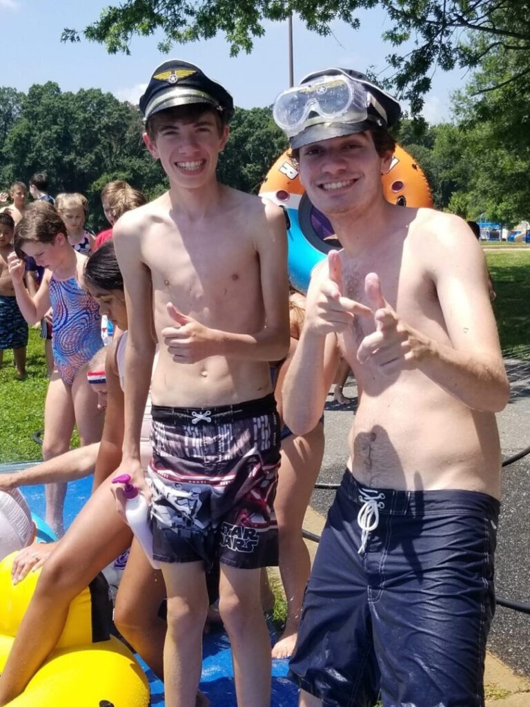 Two young men in swimwear and caps stand playfully at a water park, smiling and posing with thumbs up amidst children and inflatable rings.