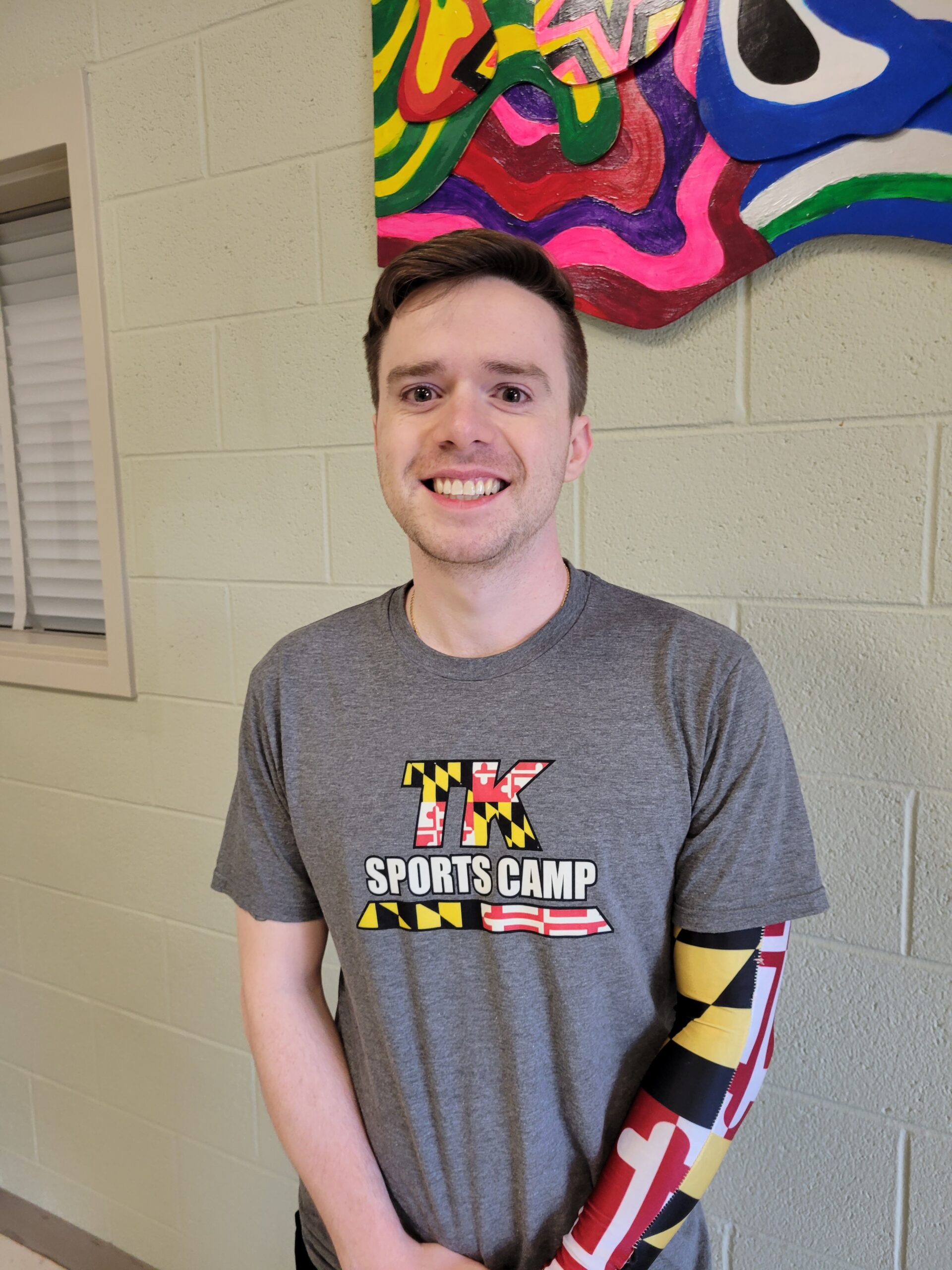 Smiling young man wearing a gray sports camp shirt and colorful arm sleeves, posing in front of a vibrant wall mural.