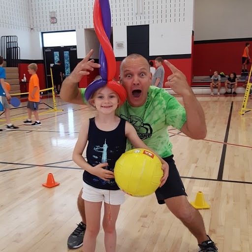 A man playfully poses with a girl holding a yellow ball, wearing a colorful balloon hat. They are in a gymnasium filled with children and activity.