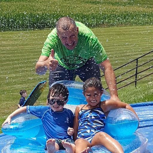 Children and an adult enjoy a sunny day on a water slide, smiling and splashing in mid-slide, surrounded by a vibrant green landscape.